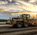 A large grader smoothing gravel at the Homer Airport with sunset colors in the sky.