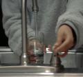 A person in a gray sweater turning on a sink faucet and filling a glass with water.