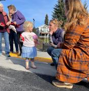 Little girl in light blue skirt holds bit of red ribbon and talks to two women kneeling besider her on sidewalk