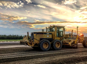 A large grader smoothing gravel at the Homer Airport with sunset colors in the sky.