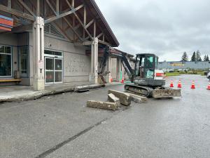 A small excavator digging curb out of the sidewalk in front of the Homer Airport terminal building under grey, cloudy skies