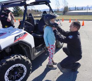 Police officer bending down to adjust helmet on girl next to a four-wheeler.