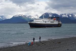 Two people on a beach watching the M/V Tustemena arriving in Homer.