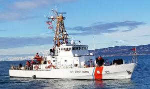 Photo of white United States Coast Guard cutter Naushon floating in still blue waters with mountains behind it.