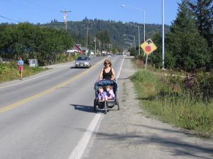 Woman walking partially on the shoulder of a road and partially on the fog line pushing a baby stroller.