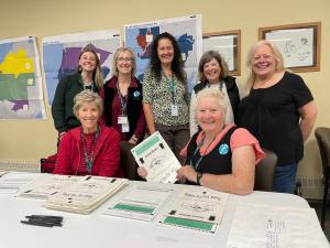 Seven women, 5 standing and 2 sitting at a white plastic table with stacks of ballot sleeves and voting precinct maps on wall.