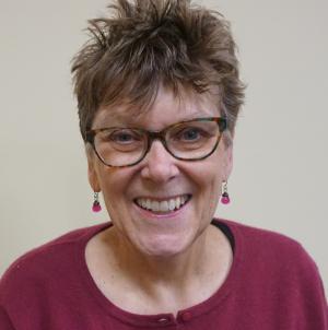 A portrait of a smiling woman with short hair and glasses wearing a raspberry-colored shirt