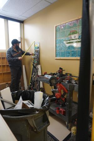 Work man in blue coveralls and cap holds yellow measuring tape up to an electronic control board leaning against a wall.