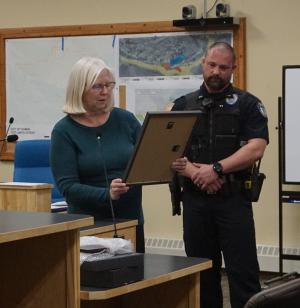 A woman in blue sweater with white hair and glasses holds a plaque and reads a citation to a police officer in uniform.