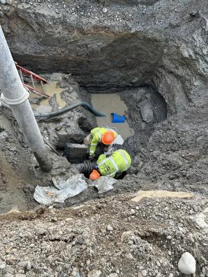 Two men in high visibility vests work in a deep muddy hole to repair a water line.
