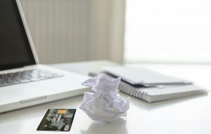 Open laptop computer on white tabletop next to credit card, crumpled piece of paper and white booklets.