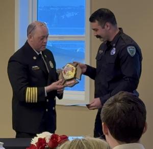 A fire chief in a blue uniform presents a wood plaque to a uniformed firefighter