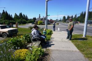 Photo of family sitting on bench and admiring a flower garden on the sidewalk at intersection of Pioneer Ave & Main St in Homer.