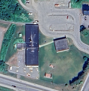 Aerial photo of a property with one large and one small building with a large parking lot on the north and a road to the south.