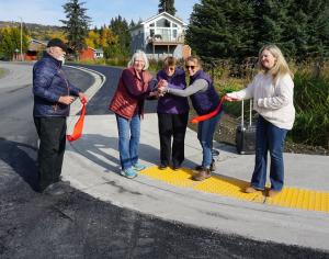 Man and woman holding ends of red ribbon, and three smiling women with big scissors cutting it in half on corner of new sidewalk