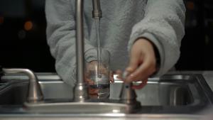 A person in a gray sweater turning on a sink faucet and filling a glass with water.