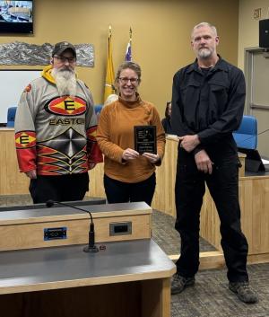 Three people standing next to each other in a City Council meeting room, two men and a woman in the middle holding an award.