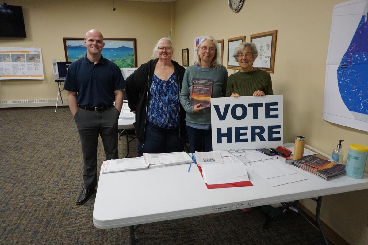 Poll workers and City Clerks stand in front of the polling place with a Vote Here sign