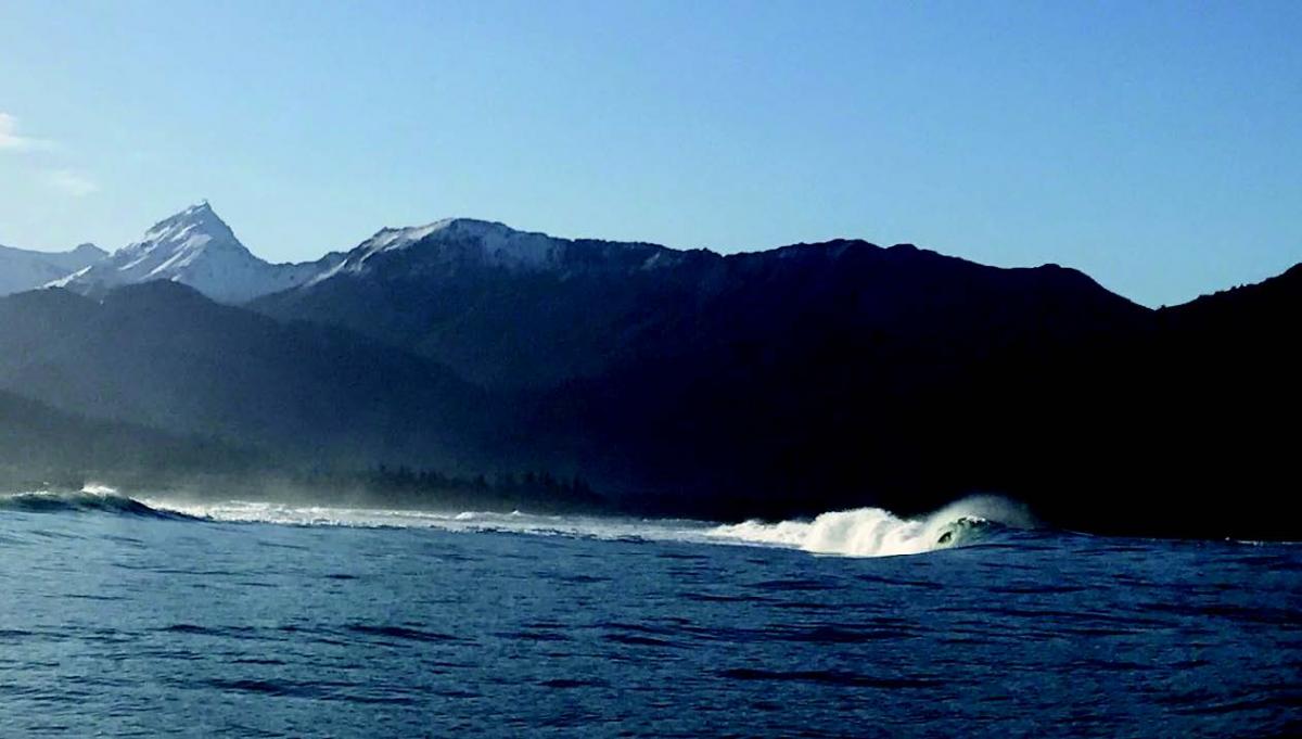 A large standing wave in an otherwise calm sea with mountains and a blue sky in the background.