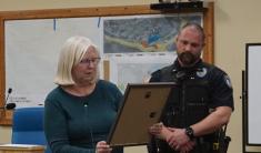 A woman in blue sweater with white hair and glasses holds a plaque and reads a citation to a police officer in uniform.