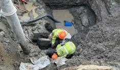 Two men in high visibility vests work in a deep muddy hole to repair a water line.