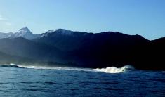 A large standing wave in an otherwise calm sea with mountains and a blue sky in the background.