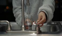 A person in a gray sweater turning on a sink faucet and filling a glass with water.